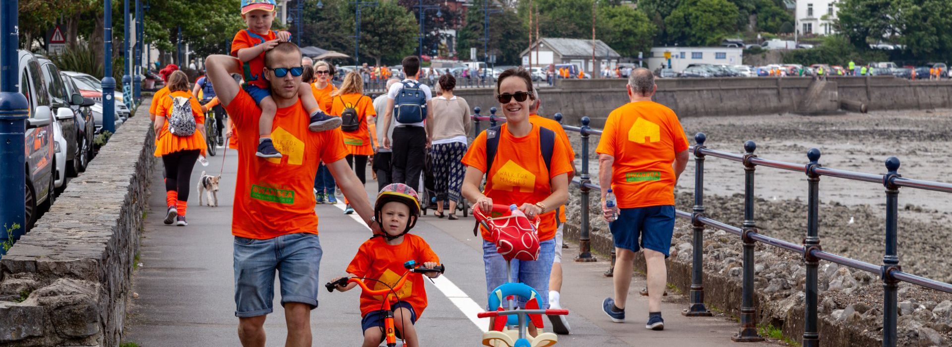 A young family wearing orange Maggie's T-shirts doing the sponsored walk, with a toddler on Dad's shoulder and a child on a bike